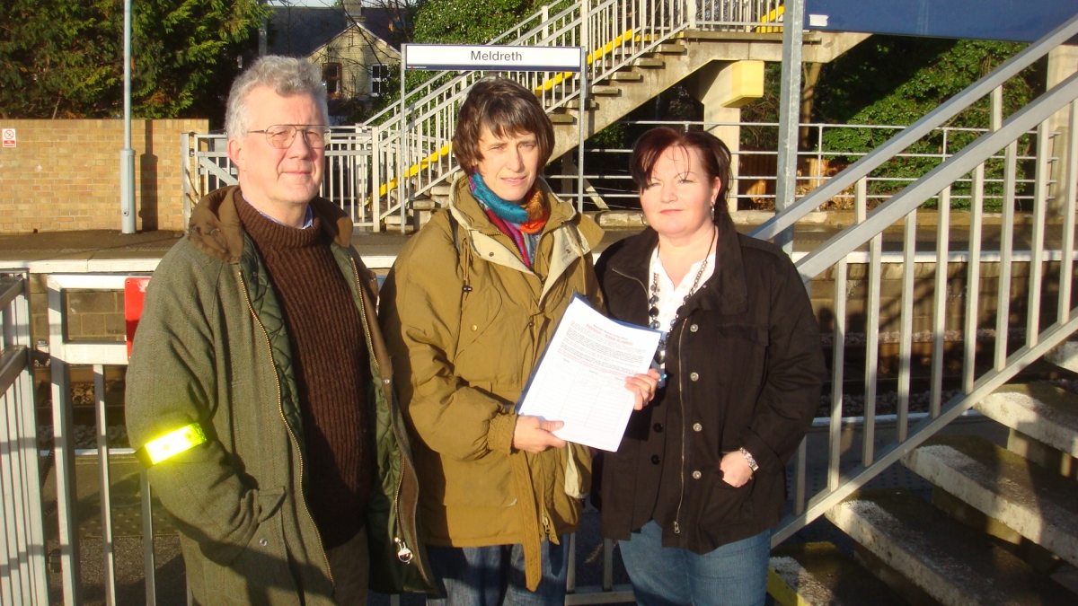 A man and two women stand in front of a sign saying 'Meldreth station' holding a paper petition 
