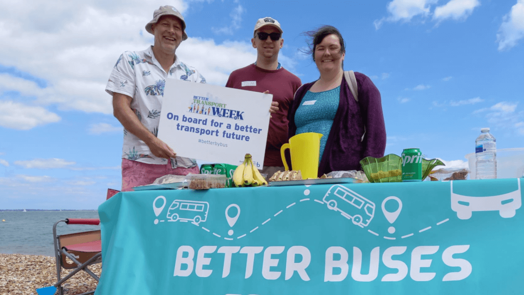 3 people standing on a beach holding a Better Transport Week sign 