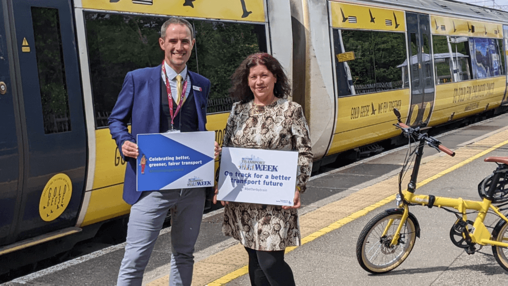 Two people at a railway station holding up a Better Transport Week sign